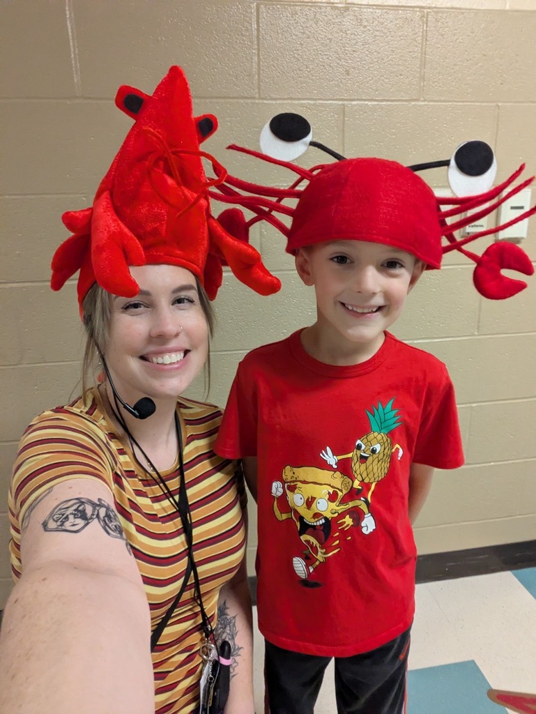 A teacher and a young student smile together in a school hallway, both wearing matching novelty lobster hats during spirit week.