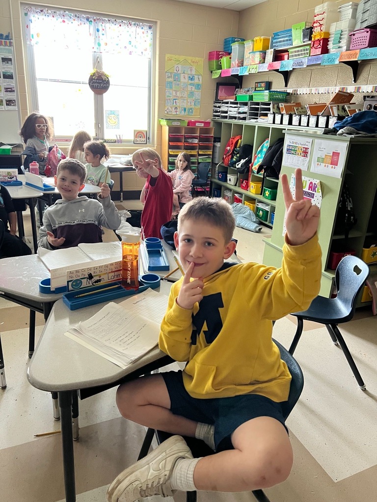 A young boy in a yellow hoodie grins and flashes a peace sign at his desk in a bright elementary classroom, with classmates visible in the background.