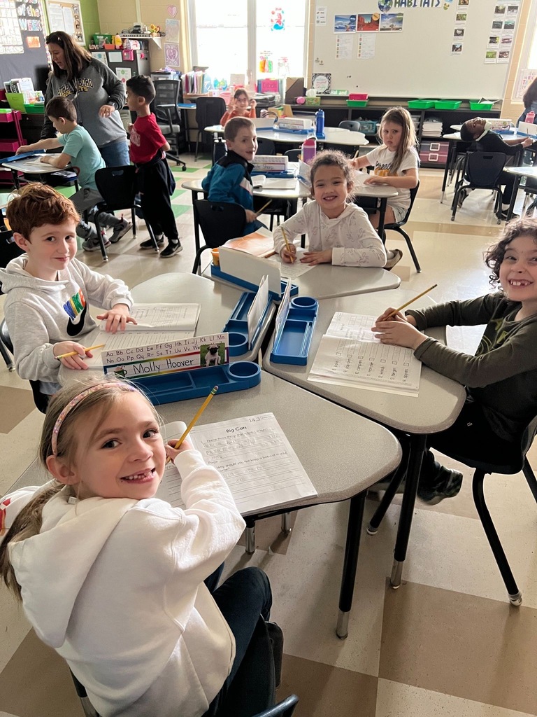 Elementary students work at cluster desks in a colorful classroom, several smiling at the camera while holding pencils and working on worksheets.