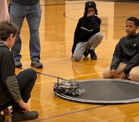 Three middle school students crouch on a gym floor, watching a small robot compete on a circular sumo arena mat.