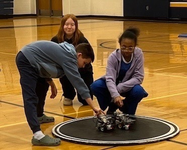 Three students kneel on a gym floor to position their robot on a circular sumo arena mat before a match.
