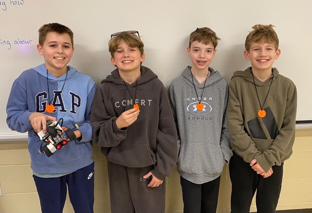 Four middle school students smile in front of a whiteboard, one holding a small LEGO robot, all wearing orange medallions around their neck