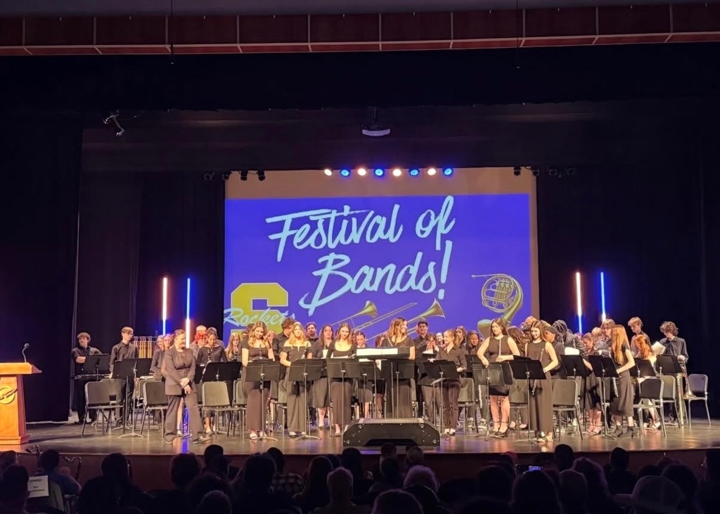 Streetsboro High School band students perform on a lit stage at the Festival of Bands, with a blue projected backdrop displaying the event name and the Rockets logo.