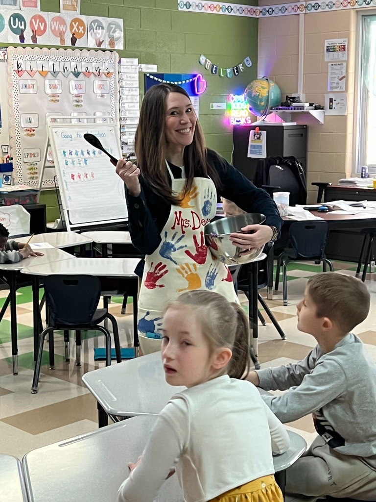 A teacher wearing a personalized handprint apron holds a mixing bowl and spoon while engaging with students in a colorful elementary classroom.