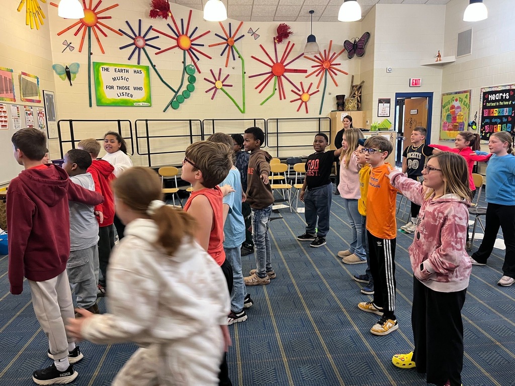 Students stand in rows in a classroom with their arms out.
