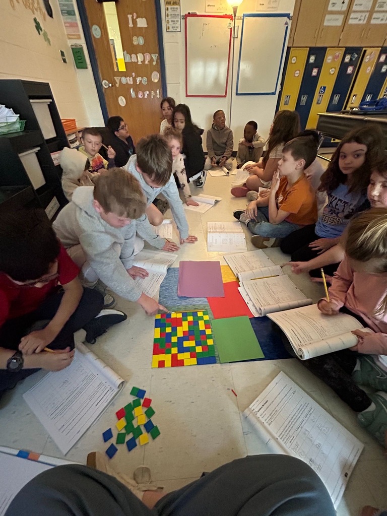 Third grade students sit in a circle on the classroom floor, working on math worksheets and manipulating colorful pattern blocks and construction paper shapes during a hands-on math lesson.