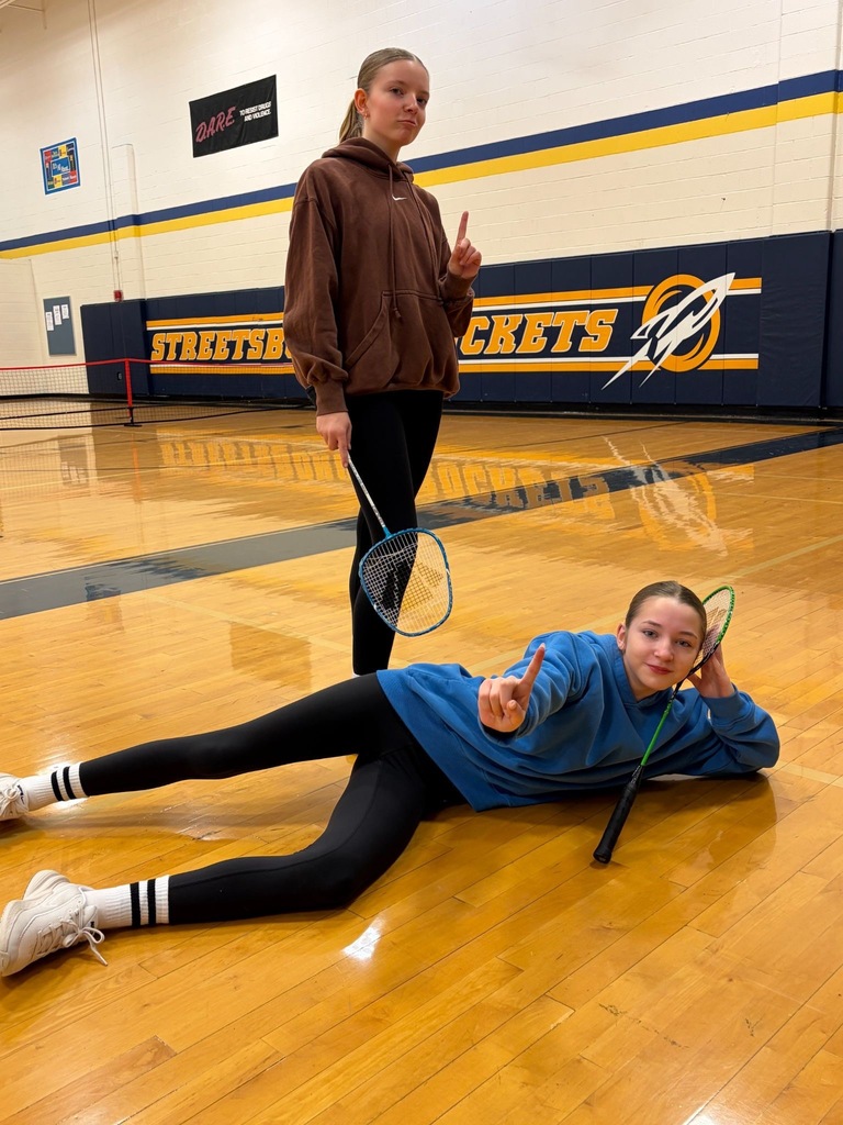One student standing holding a badminton racket, one laying on the floor of the gym holding the racket.