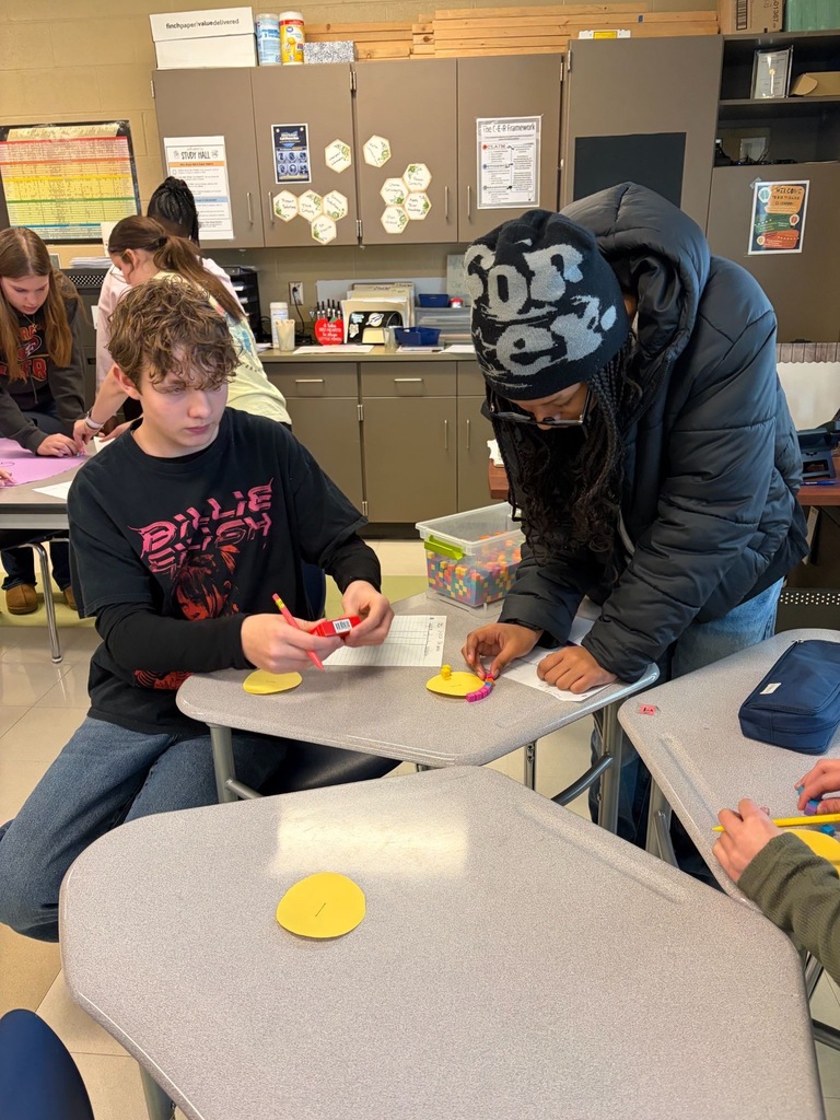 Two middle school students work together at their desks with small colorful manipulatives and a recording sheet during a hands-on math activity.