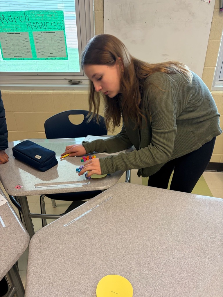 A student works with small colorful manipulatives at her desk, with a March Madness bracket poster visible on the projected screen in the background.