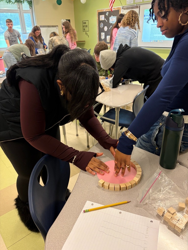 1:07 PMTwo students arrange small wooden blocks around the edge of a pink circular cutout on a desk during a hands-on Pi Day math activity, with other students working in the background.