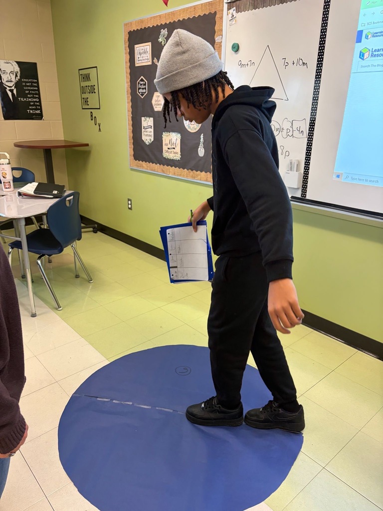 A student stands on a large blue circle on the classroom floor, holding a clipboard and recording data during a hands-on Pi Day math activity.