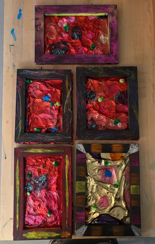 Five student-created abstract landscape paintings displayed on a wooden surface, featuring textured mixed-media compositions with crinkled metallic materials, bold red and jewel-toned colors, and small green embellishments, each in a painted frame.