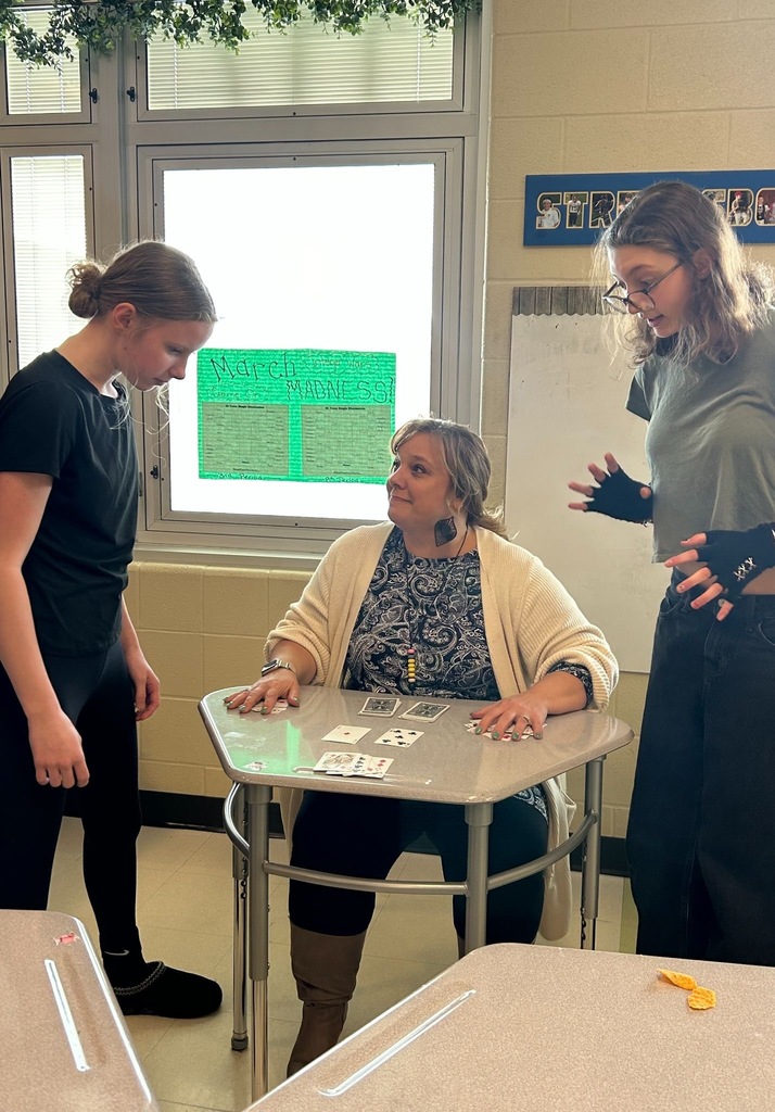 A teacher sits at a student desk with playing cards spread in front of her while two students stand on either side, all focused on the cards. A "March Madness" tournament bracket display is visible on the window behind them, and a "Streetsboro" sign is on the wall.