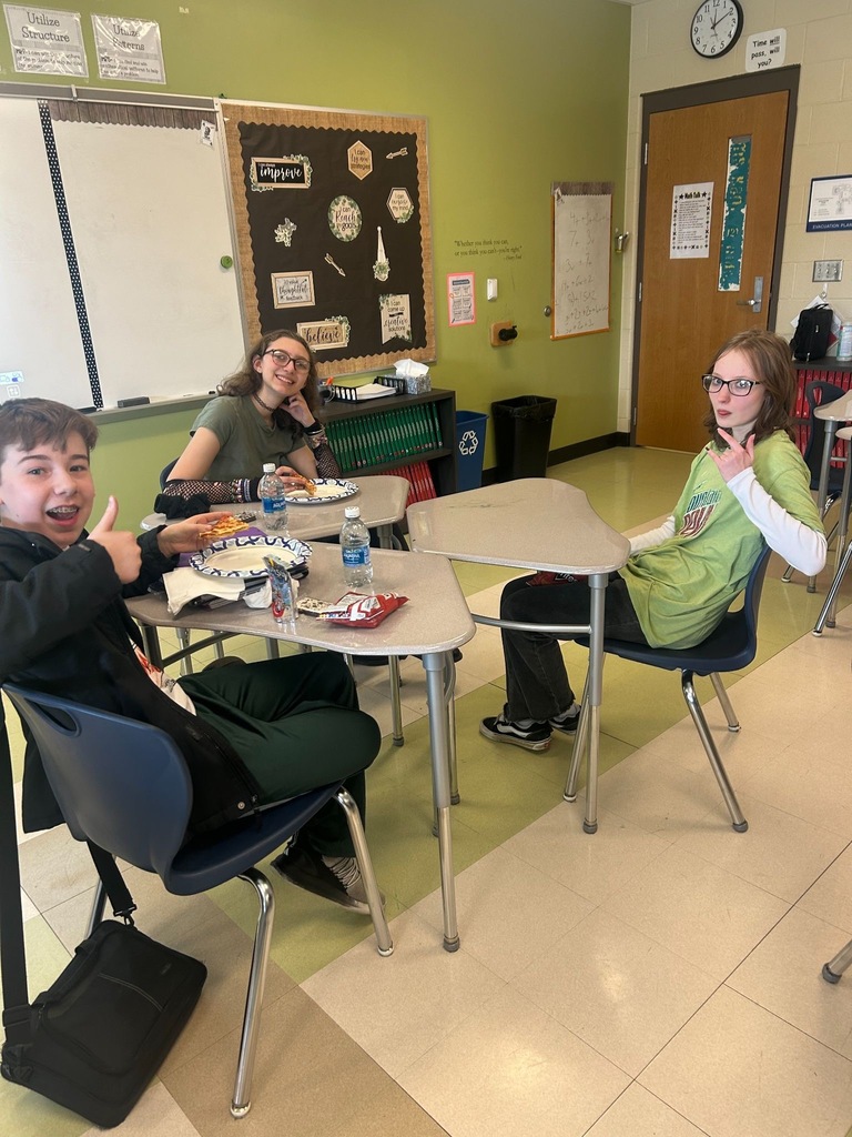 Three middle school students sit at pushed-together desks in a classroom, eating lunch and posing for the camera. One student gives a thumbs up, another smiles with their chin resting on their hand, and a third flashes a hand sign. Pizza, chips, and water bottles are visible on the desks.