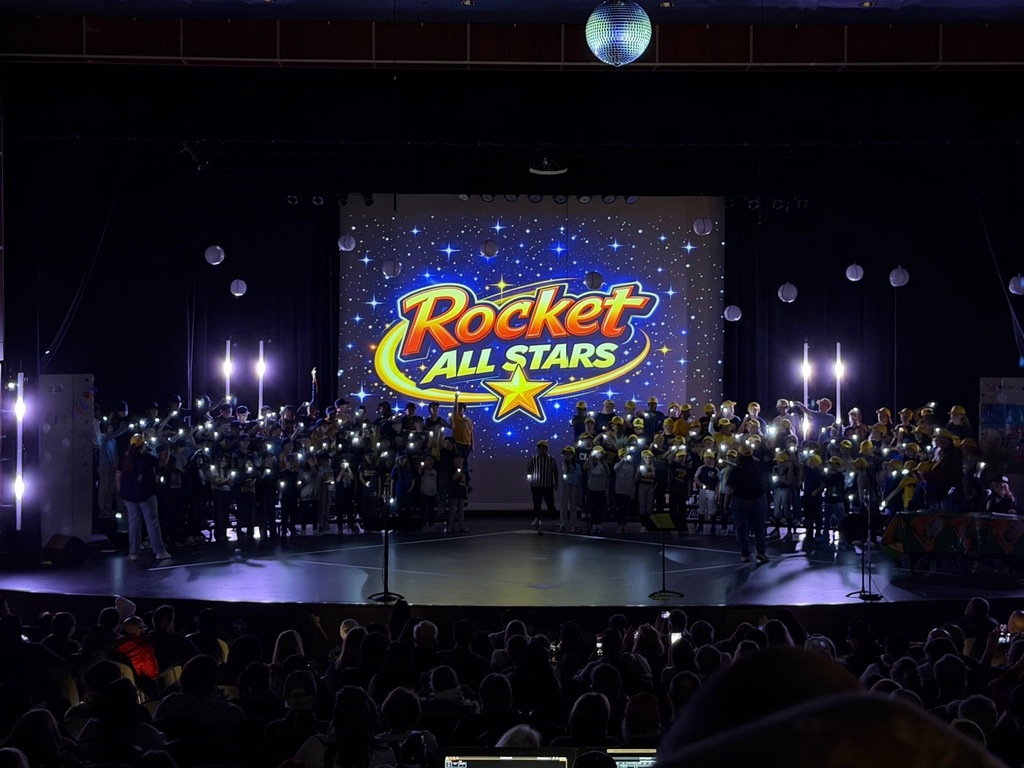 The full cast of third-grade performers holds up glowing lights on a darkened stage beneath a projected "Rocket All Stars" logo surrounded by stars, creating a dramatic finale moment as the audience watches from below.
