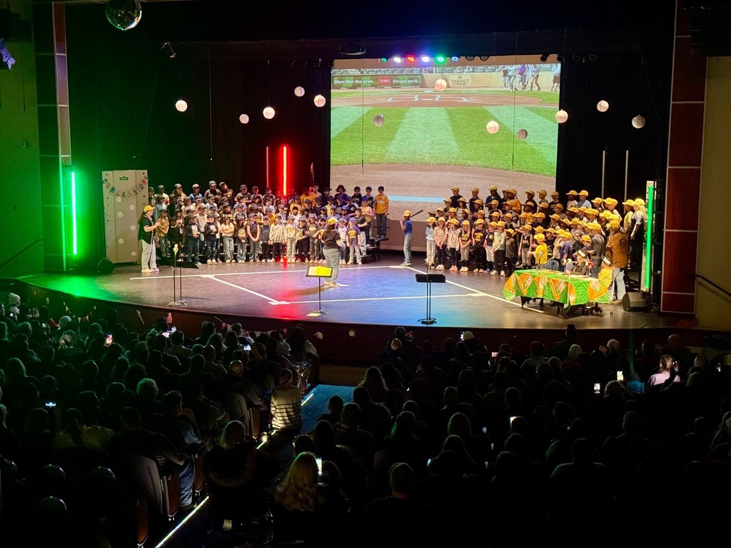 Hundreds of third-grade students in yellow baseball caps fill the stage of a large auditorium for their music concert, with a baseball field projected on the screen behind them and colorful stage lighting illuminating the performance. Families and friends pack the seats below, many capturing the moment on their phones.