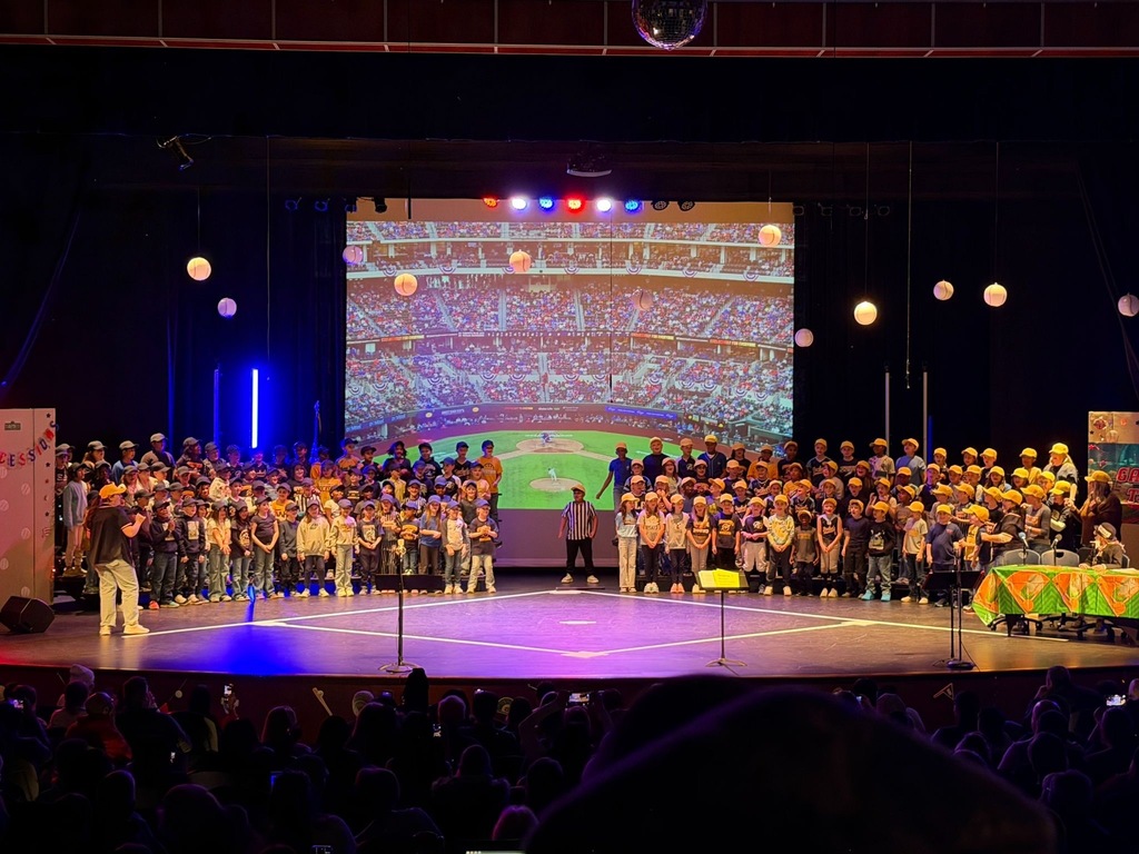 Third-grade students line the stage in matching yellow baseball caps during their spring music concert, with a projected baseball stadium backdrop behind them and globe-shaped decorations hanging overhead. A packed auditorium of families looks on as a director leads the performance.