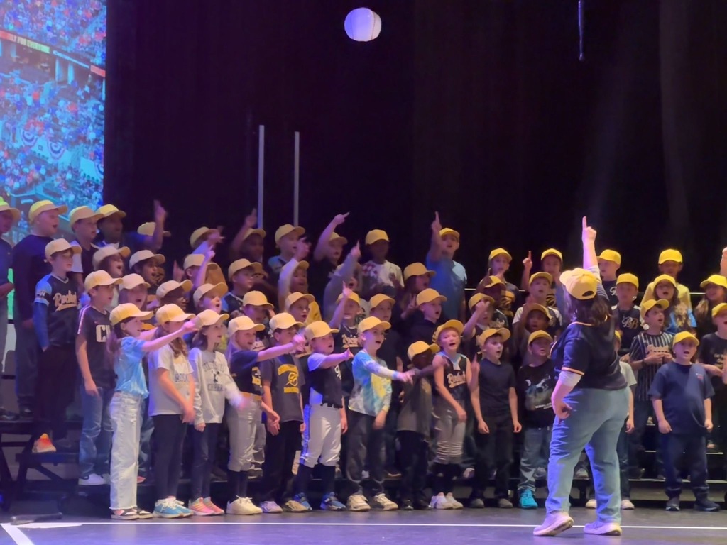 A close-up of third-grade students in yellow baseball caps singing enthusiastically on stage, many pointing upward in unison during a lively number. A director faces the group from the front of the stage as a baseball stadium projection glows in the background.