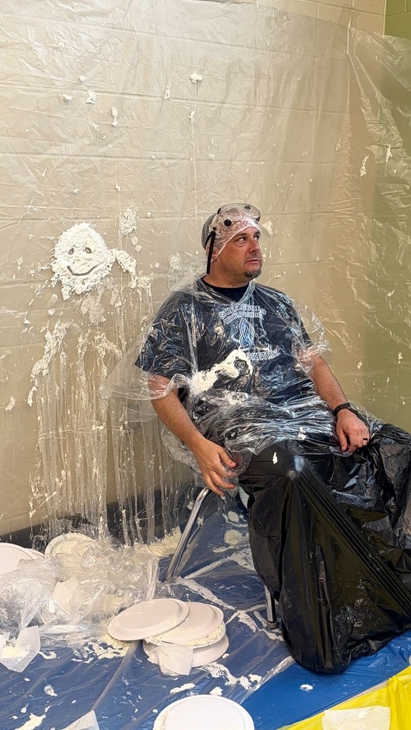 A staff member sits covered in whipped cream after being pied by students during a Pi Day celebration, wearing protective goggles and wrapped in plastic sheeting. Empty pie plates litter the floor around him, and a whipped cream smiley face is visible on the plastic backdrop behind him.