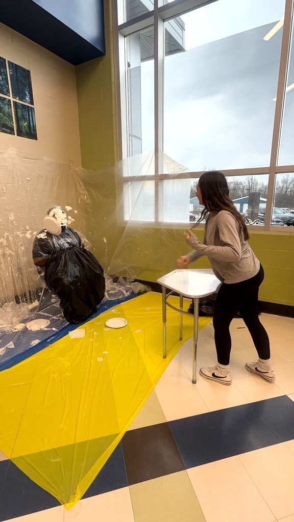 A middle school student winds up to throw a pie at a staff member during a Pi Day fundraiser event. The staff member is seated and wrapped in a black trash bag for protection, with yellow and blue plastic sheeting covering the floor.