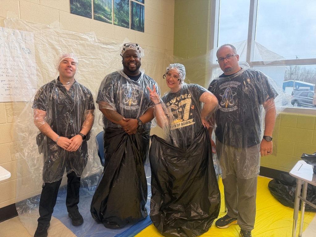 : Four Streetsboro Middle School staff members pose together before their turn in the pie booth, dressed in matching school shirts, clear plastic ponchos, hair nets, and black trash bags. One flashes a peace sign while another smiles broadly, all good-naturedly prepared for the messy Pi Day fun.