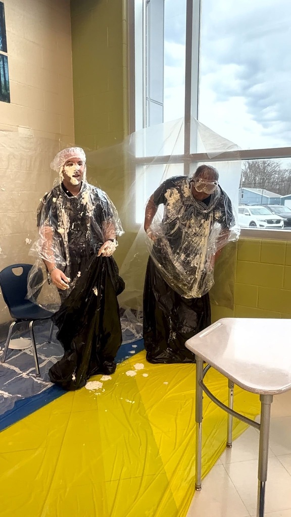 Two whipped-cream-covered staff members stand side by side after being pied by students, both wearing protective goggles, hair nets, plastic ponchos, and black trash bags. They laugh and survey the mess on the yellow-covered floor around them.