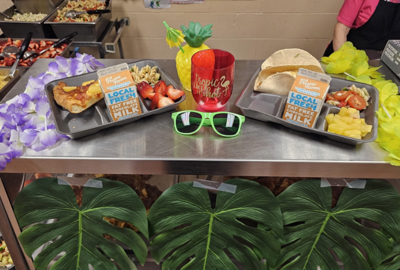 Two school lunch trays display the beach-themed menu at Streetsboro Elementary School, featuring Hawaiian pizza, fish tacos, fresh strawberries, pasta salad, tropical fruit, and Prairie Farms chocolate milk cartons. The trays are surrounded by tropical decorations including a pineapple cup, lei flowers, green sunglasses, and large palm leaves.