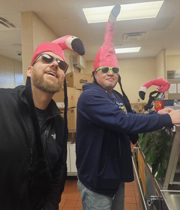 Two school cafeteria staff members wear novelty pink flamingo hats and sunglasses while serving lunch during a beach-themed meal event at Streetsboro Elementary School. Both are grinning as they work the serving line.