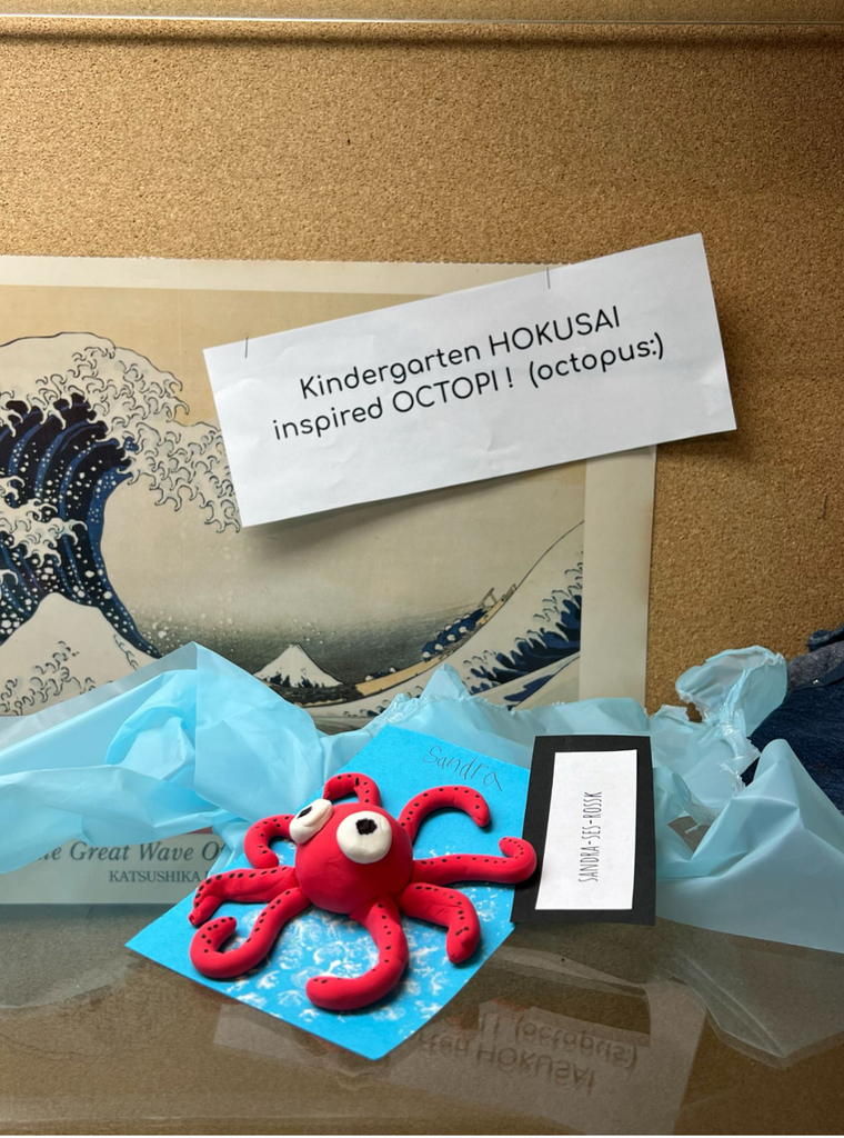 A red clay octopus sculpture sits on a blue paper "ocean" at a school art display. Behind it, a print of Hokusai's The Great Wave off Kanagawa is pinned to a corkboard alongside a sign reading "Kindergarten HOKUSAI inspired OCTOPI! (octopus:)". A name card reads "Sandra Ses-Rossk."