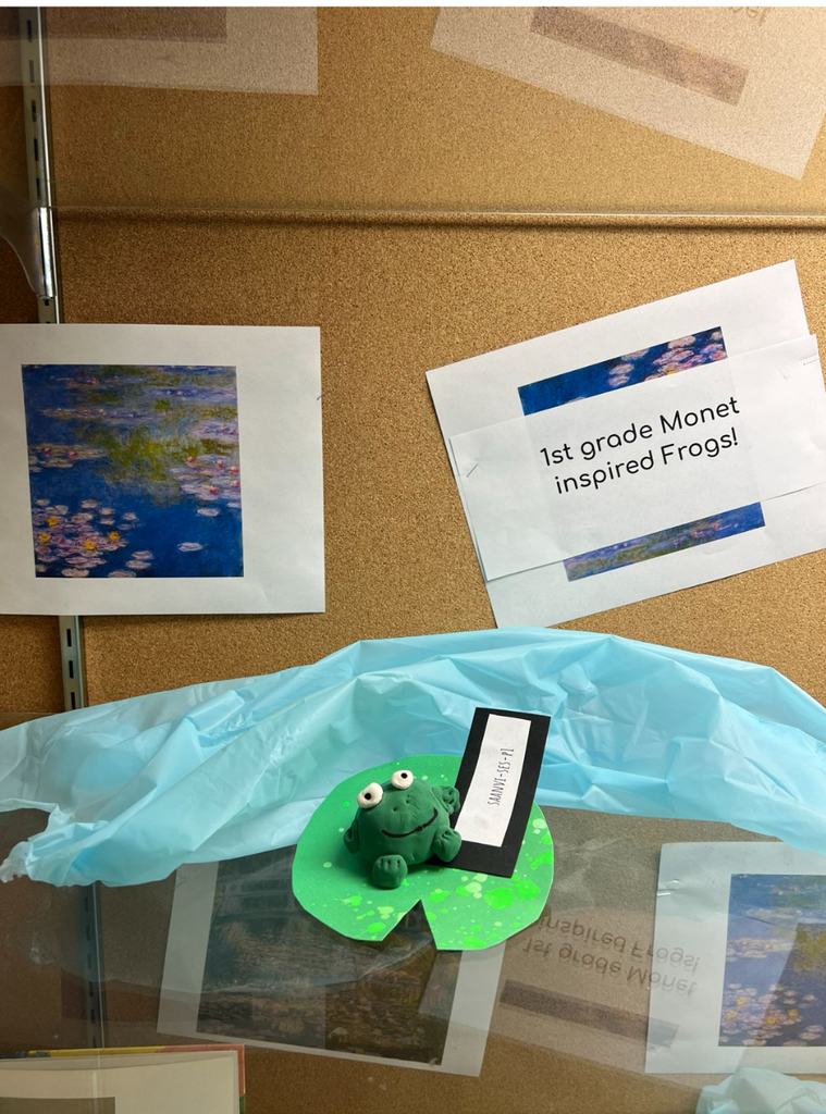 A green clay frog sculpture sits on a paper lily pad at a school art display. Behind it, a print of Monet's Water Lilies is pinned to a corkboard alongside a sign reading "1st grade Monet inspired Frogs!" Blue tissue paper suggests water around the sculpture.
