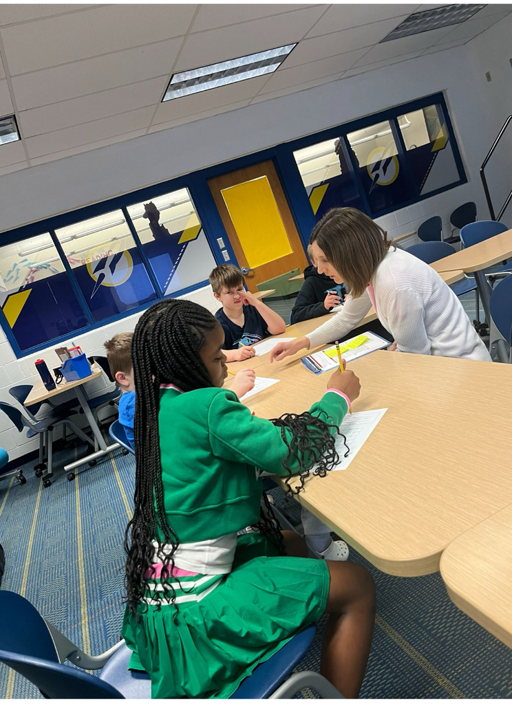 9:03 AMA teacher leans over a kidney-shaped small-group table to assist a student, while two other elementary-age students work on paper worksheets with pencils. The classroom features blue and yellow decor with large windows. A fourth student works independently at a separate desk in the background.