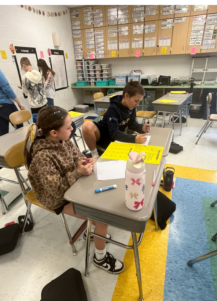 Two upper elementary or middle school students work at pushed-together desks during a math activity, with a yellow "Four in a Row" division game board on the desk between them. In the background, other students work at the wall-mounted Wipebook whiteboards. The classroom walls feature extensive math vocabulary and fraction reference materials on upper cabinets.
