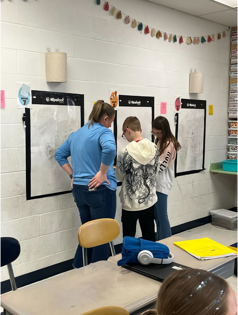  A teacher in a blue top works alongside two middle school students at a "Group 8" Wipebook reusable whiteboard mounted on a classroom wall. Two additional numbered group whiteboards are visible on either side. A tassel garland decorates the wall above.