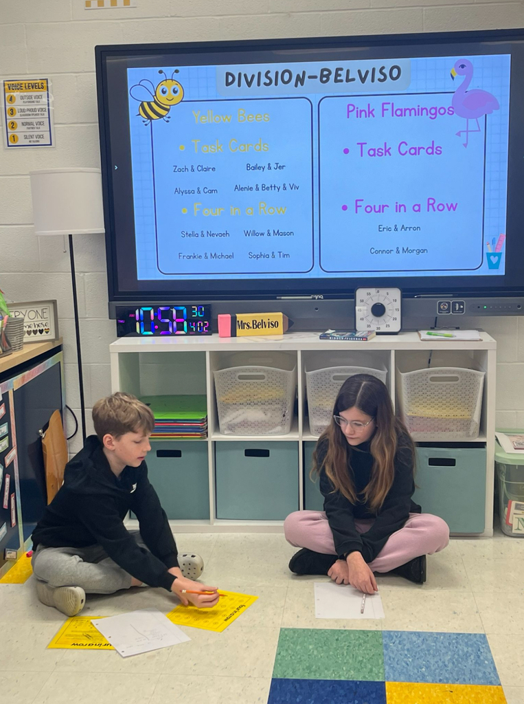 Two elementary students sit cross-legged on the classroom floor playing a division math game. A large display screen behind them shows a "Division – Belviso" slide listing student pairs assigned to "Yellow Bees" and "Pink Flamingos" groups for Task Cards and Four in a Row activities. A yellow game board and dice are visible on the floor between them.