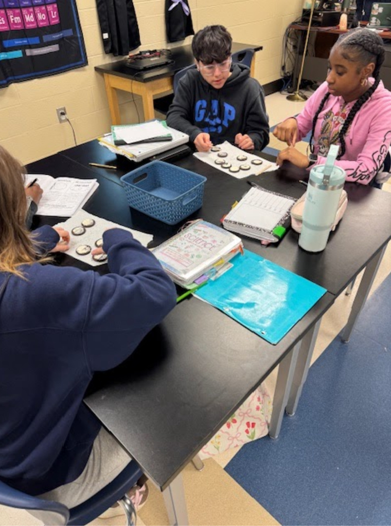  Three middle school students work together at a black lab table in a science classroom, each manipulating Oreo cookies on worksheets — a common activity for modeling moon phases. A periodic table is visible on the wall in the background. Student science notebooks and binders are spread across the table.
