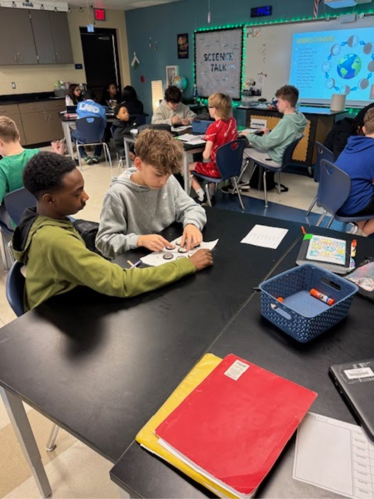 A wide view of a middle school science classroom where students at multiple lab tables use Oreo cookies to model moon phases on worksheets. A "Science Talk" bulletin board and a projected slide showing moon phase diagrams with a globe illustration are visible at the front of the room. Supply baskets with markers sit on the tables.