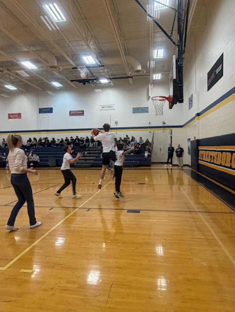 A player in a white shirt leaps toward the basket for a layup attempt during a basketball game in the Streetsboro school gymnasium. Teammates and an opposing player react nearby, while a crowd fills the bleachers along the wall.