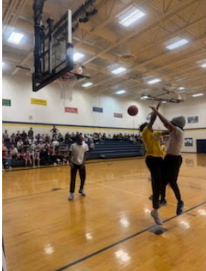 A player in a yellow shirt goes up for a shot while being defended during a basketball game in a school gymnasium. Packed bleachers of spectators watch in the background.
