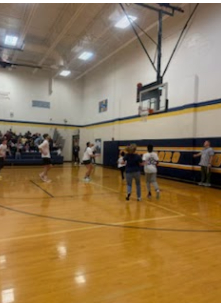 A wide-angle view of ongoing play during a school basketball game in the Streetsboro gymnasium. Several players in white shirts move across the court, with a large crowd of spectators visible along the sideline bleachers.