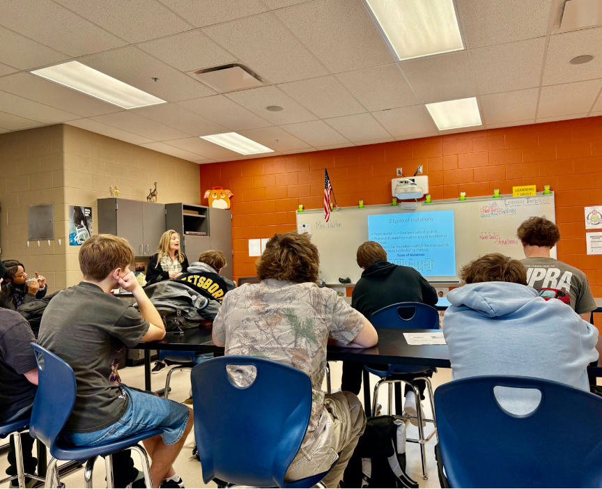 A teacher addresses a class of high school students from the front of a room with an orange accent wall. A projected slide titled "3 Types of Mutations" is visible on the whiteboard behind her. Upcoming test reminders for Anatomy and Biology are posted on the side wall.
