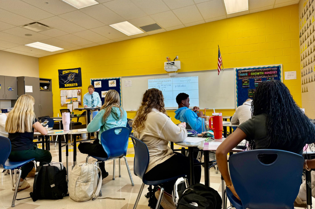 A male teacher in a blue shirt and tie instructs a high school math class in a bright yellow-walled classroom. A projected whiteboard displays notes on graphing rational functions. A Streetsboro Rockets banner and a "I Have a Growth Mindset" bulletin board are visible on the walls.