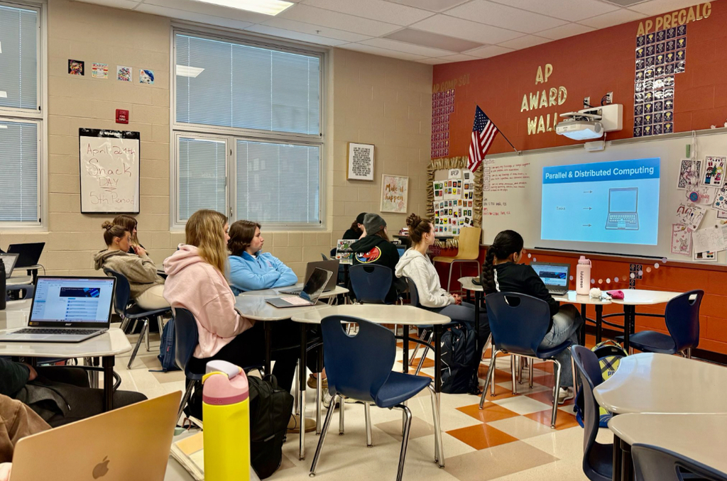 High school students with laptops face a projected slide on "Parallel & Distributed Computing" in a classroom decorated with an orange "AP Award Wall" and "AP Precalc" signage. A whiteboard notes "April 29th Snack Day 5th Period."