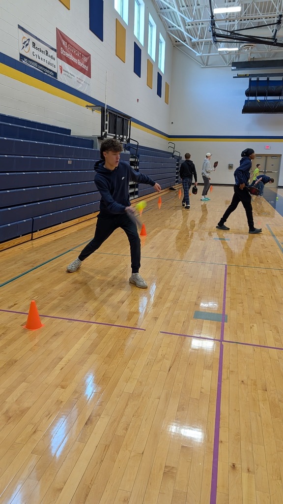 Here's alt text for each of the three photos: Photo 1 (left): "A student in a black Nike outfit plays pickleball in a school gymnasium, mid-swing, with orange cones marking the court and other students visible in the background.