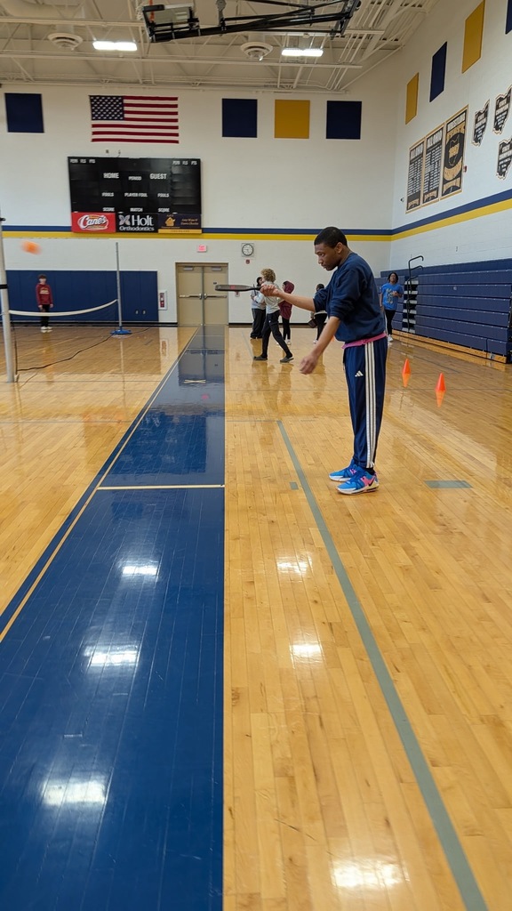  student in a navy Adidas tracksuit prepares to serve or toss a ball in a school gymnasium set up for pickleball, with orange cones, a net, and other students playing in the background beneath an American flag.