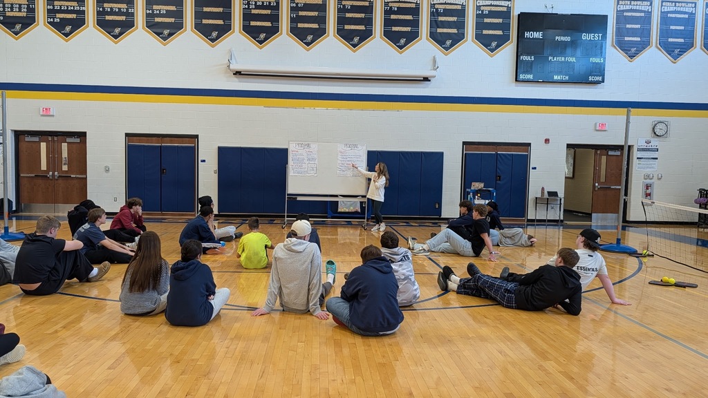 A large group of middle school students sit on the gymnasium floor in rows, attentively facing a teacher or instructor standing near a whiteboard at the far end of the gym.