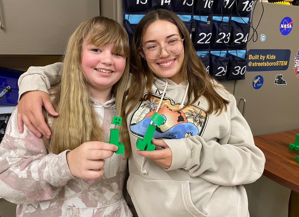 Two smiling middle school students pose together, each holding a green 3D-printed Minecraft bobblehead they created in STEM class.