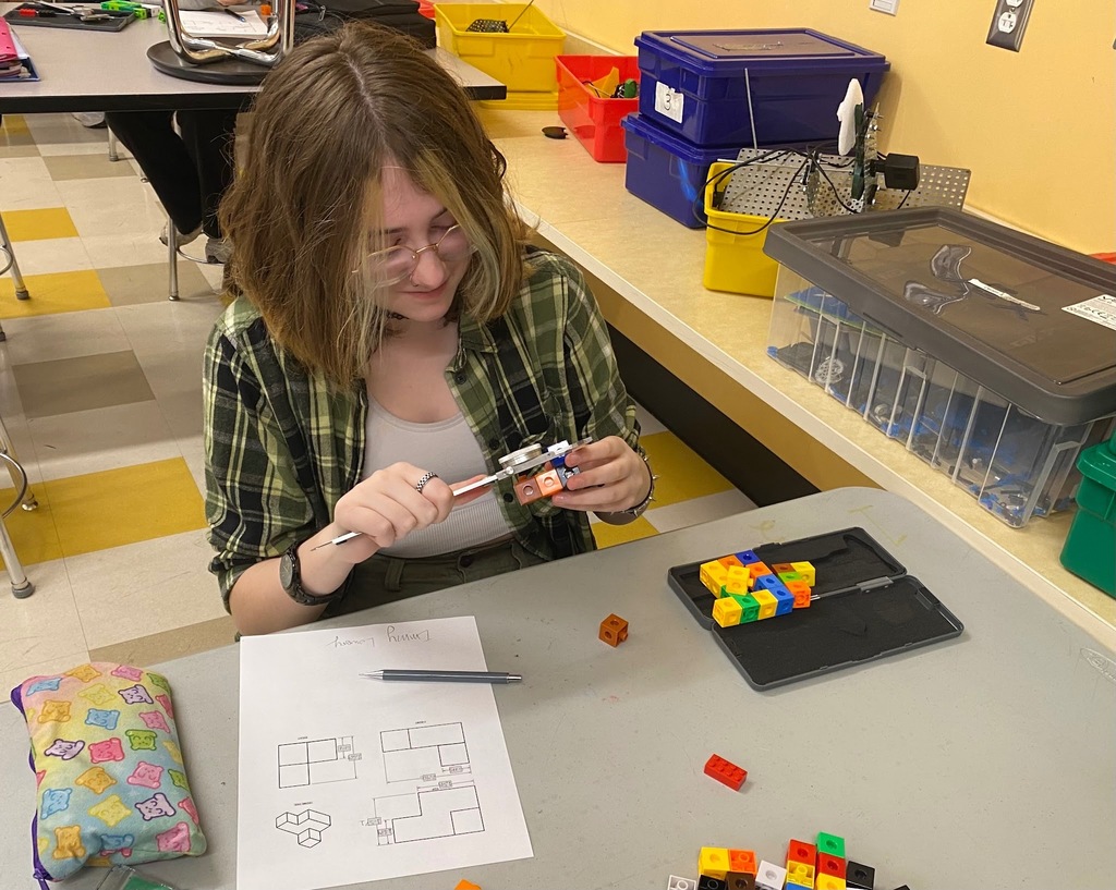 A middle school student uses a dial caliper to carefully measure a small object at her desk, with colorful building materials and a technical drawing visible nearby