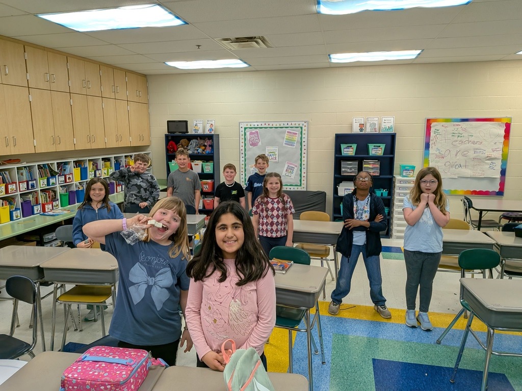 Elementary students sit at their desks eating lunch in a colorful classroom, smiling at the camera, with bookshelves and a bulletin board visible in the background