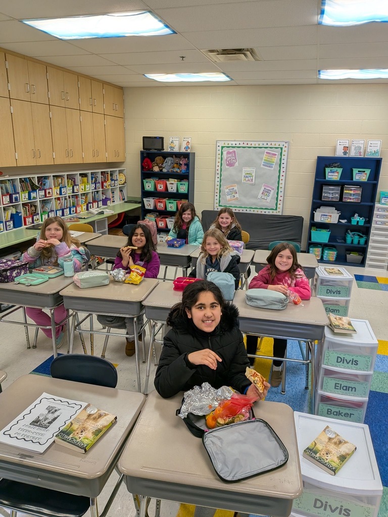Elementary students sit at their desks eating lunch in a colorful classroom, smiling at the camera, with bookshelves and a bulletin board visible in the background.