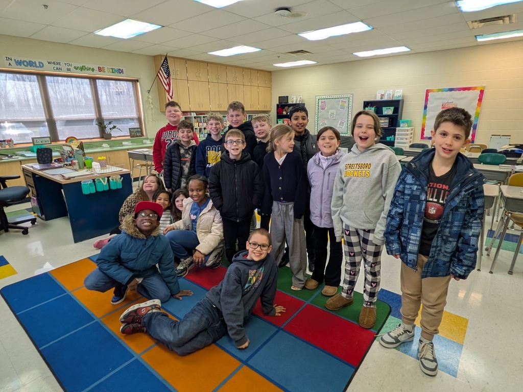 A large group of elementary students pose together for a class photo in their classroom, some sitting on a colorful rug and others standing, with an American flag and cheerful wall decorations in the background.